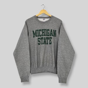 Puede incluir: Una sudadera gris de cuello redondo con las palabras "MICHIGAN STATE" impresas en letras verdes. La sudadera está colgada de una percha de madera sobre un fondo claro. La sudadera tiene mangas largas y un cuello acanalado.