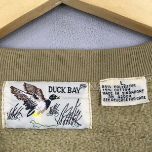 May include: Close-up of a tan sweatshirt collar with a "DUCK BAY" label featuring a duck graphic. The label also includes text about the fabric content and origin. A size "L" tag is visible.