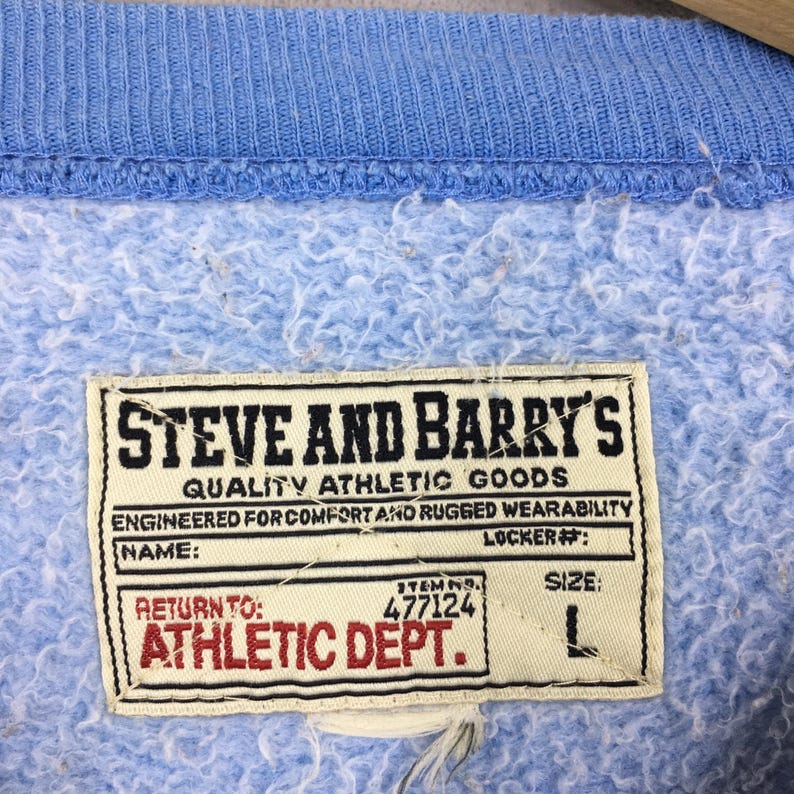 May include: Close-up of a pale blue sweatshirt with a ribbed collar. The label reads "STEVE AND BARRY'S QUALITY ATHLETIC GOODS" with the text "ATHLETIC DEPT." and size "L".