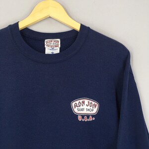 May include: A navy blue sweatshirt featuring the "Ron Jon Surf Shop U.S.A." logo in a white and red oval. The garment is suspended on a wooden hanger, showcasing its design and colour.