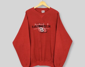 May include: A red sweatshirt with the text "DALE EARNHARDT JR" above a red and white number "8" logo. The sweatshirt is hanging on a wooden hanger against a light gray background. The sweatshirt is a crew neck style.