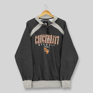 May include: A charcoal gray Cincinnati Bengals sweatshirt with a gray collar and accents. The team name and logo are printed on the front in orange, black, and white. The sweatshirt has a quarter-zip closure and a gray ribbed hem and cuffs.
