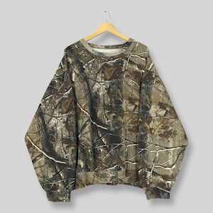 Op de afbeelding: Een sweatshirt met camouflagepatroon in bruin-, groen- en beigetinten, hangend aan een houten hanger. De top met lange mouwen heeft een ronde hals en geribde manchetten, met een dicht bosachtig ontwerp.