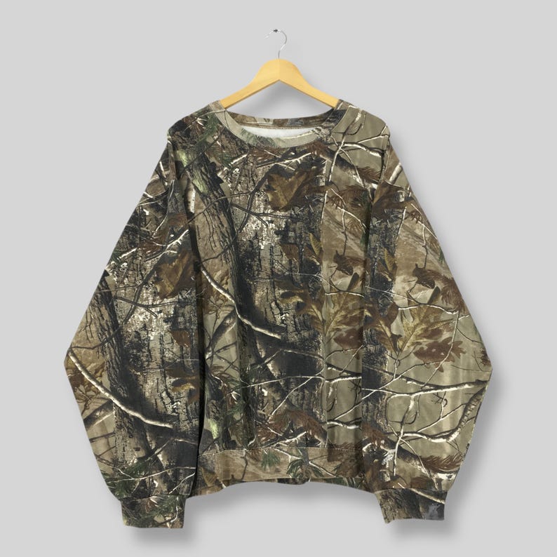 Op de afbeelding: Een sweatshirt met camouflagepatroon in bruin-, groen- en beigetinten, hangend aan een houten hanger. De top met lange mouwen heeft een ronde hals en geribde manchetten, met een dicht bosachtig ontwerp.