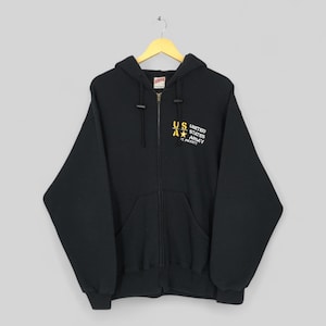 May include: Black zip-up hooded sweatshirt with a front pocket and drawstring hood. The sweatshirt features the text "US ARMY" in yellow and white on the upper left chest. The sweatshirt is hanging on a wooden hanger.