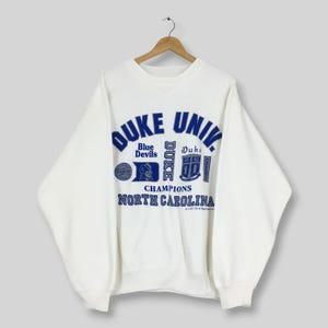 Może przedstawiać: Biała bluza z długim rękawem i okrągłym dekoltem z niebieskimi nadrukami przedstawiającymi napisy DUKE UNIV., Blue Devils, Duke oraz North Carolina. Wzór zawiera logo tarczy, profil maskotki oraz tekst 1992 CD All Rights Reserved.