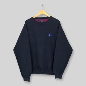 Könnte beinhalten: Marineblaues Sweatshirt mit einem kleinen gestickten Design auf der Brust. Das Sweatshirt hat einen Rundhalsausschnitt mit einem bordeauxfarbenen Kragen und einem kleinen gestickten Logo. Das langärmlige Oberteil hängt an einem Holzbügel.
