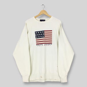May include: Cream-colored sweatshirt featuring a graphic of the American flag in navy blue, red, and white. The text "RALPH LAUREN POLO SPORT" is printed below the flag. The long-sleeved top is hanging on a wooden hanger.