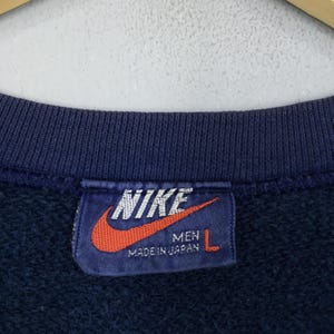 May include: Navy blue sweatshirt with a tag featuring the Nike logo in white and a red swoosh. The tag reads "MEN" and "MADE IN JAPAN" with a size "L". The sweatshirt is a vintage style.