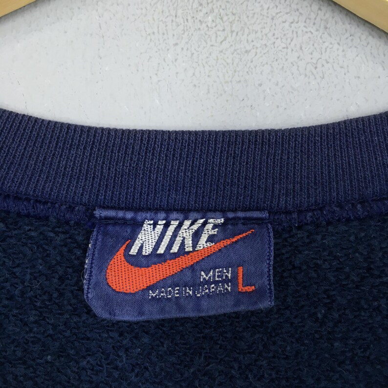May include: Navy blue sweatshirt with a tag featuring the Nike logo in white and a red swoosh. The tag reads "MEN" and "MADE IN JAPAN" with a size "L". The sweatshirt is a vintage style.