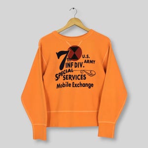 May include: A bright orange long-sleeve sweatshirt featuring a black graphic print that reads 7th INF DIV. U.S. ARMY SPECIAL SERVICES Mobile Exchange, with a circular red and black emblem above the text and a ribbed crew neckline and cuffs.