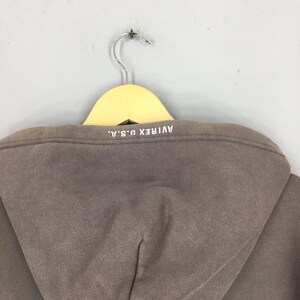 Puede incluir: Una sudadera con capucha marr&oacute;n con el texto "AVIREX U.S.A." impreso en blanco en la parte posterior de la capucha. La sudadera est&aacute; colgada de una percha de madera.