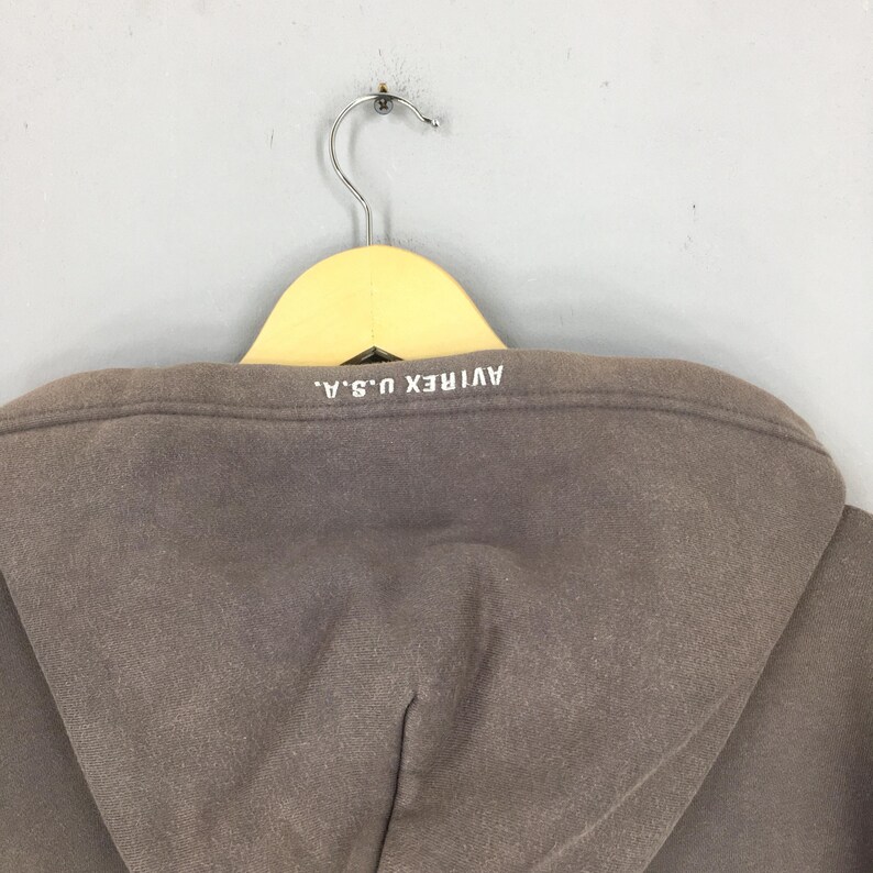 Puede incluir: Una sudadera con capucha marr&oacute;n con el texto "AVIREX U.S.A." impreso en blanco en la parte posterior de la capucha. La sudadera est&aacute; colgada de una percha de madera.