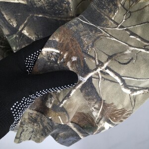 Op de afbeelding: Close-up van een camouflagepatroonstof in tinten groen, bruin en bruin. De stof wordt vastgehouden door een zwarte handschoen met witte stippen op de palm.