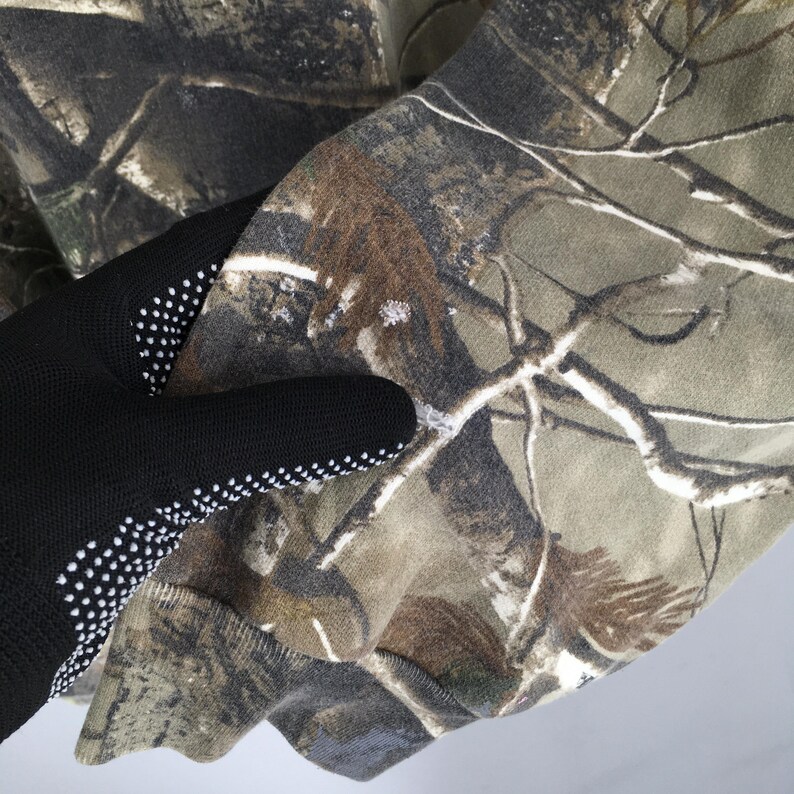 Op de afbeelding: Close-up van een camouflagepatroonstof in tinten groen, bruin en bruin. De stof wordt vastgehouden door een zwarte handschoen met witte stippen op de palm.
