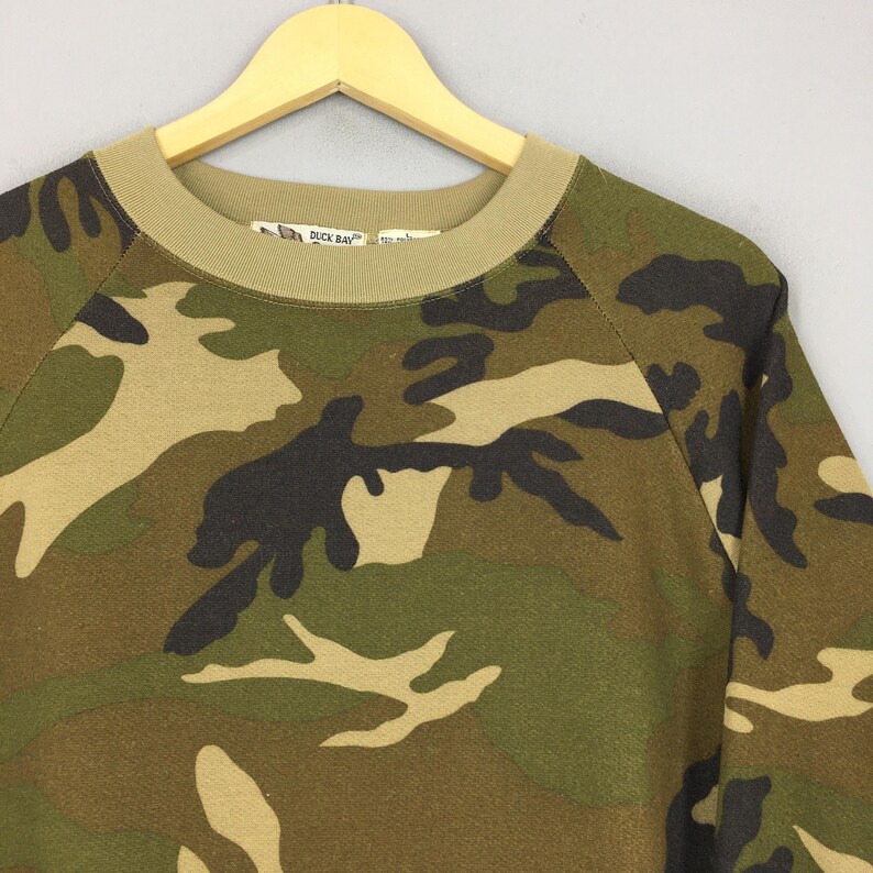 May include: Camouflage-patterned sweatshirt in shades of green, brown, and black. The crew neck and cuffs are a solid tan color. The brand name "DUCK BAY" is visible on the label. The sweatshirt is hanging on a wooden hanger.