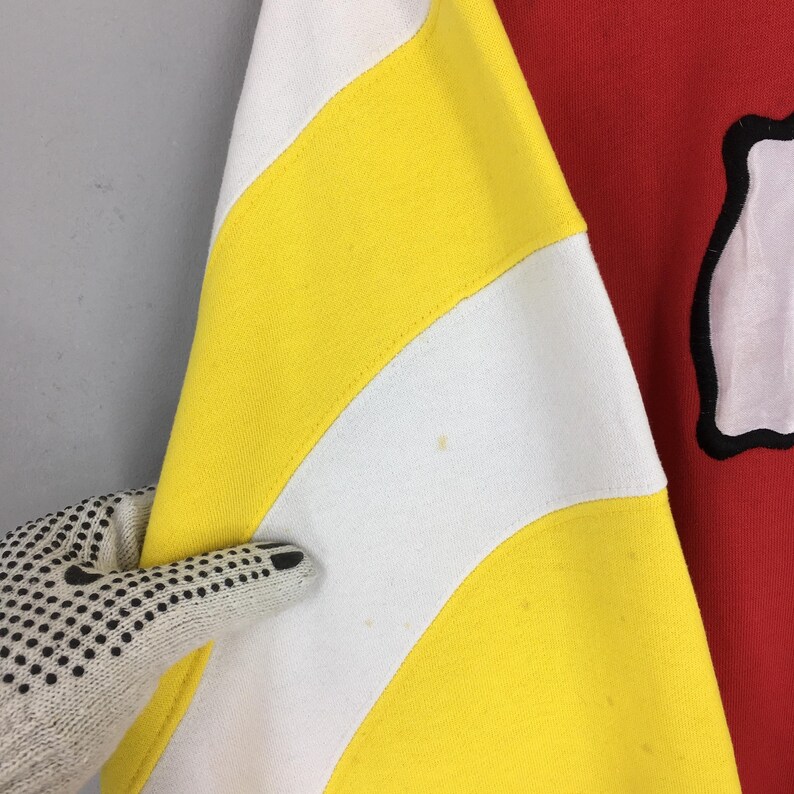 May include: Close-up of a red and yellow striped sweatshirt. The sweatshirt has a white and yellow striped pattern on the sleeve. The other side of the sweatshirt is red with a white graphic.