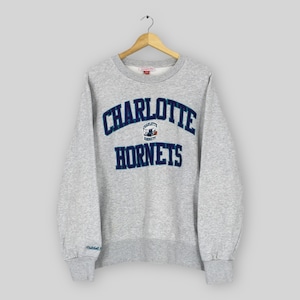 May include: A light gray sweatshirt with the words "CHARLOTTE HORNETS" in dark blue and teal lettering. The sweatshirt has a crew neck and long sleeves. The Mitchell & Ness logo is visible.