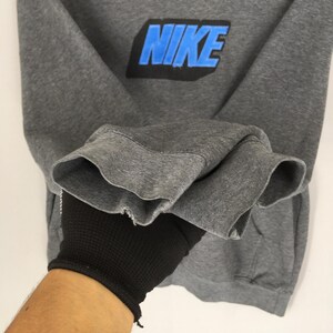 Może przedstawiać: Szara bluza z napisem "NIKE" w niebieskich i czarnych literach na piersi. Widoczne są mankiety rękaw&oacute;w. Odzież trzymana jest przez dłoń w rękawicy.