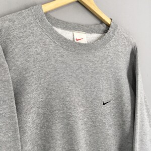 May include: A gray crew neck sweatshirt with a small black Nike swoosh logo on the chest. The sweatshirt has a ribbed neckline and cuffs. The Nike logo is also visible on the tag inside the collar.