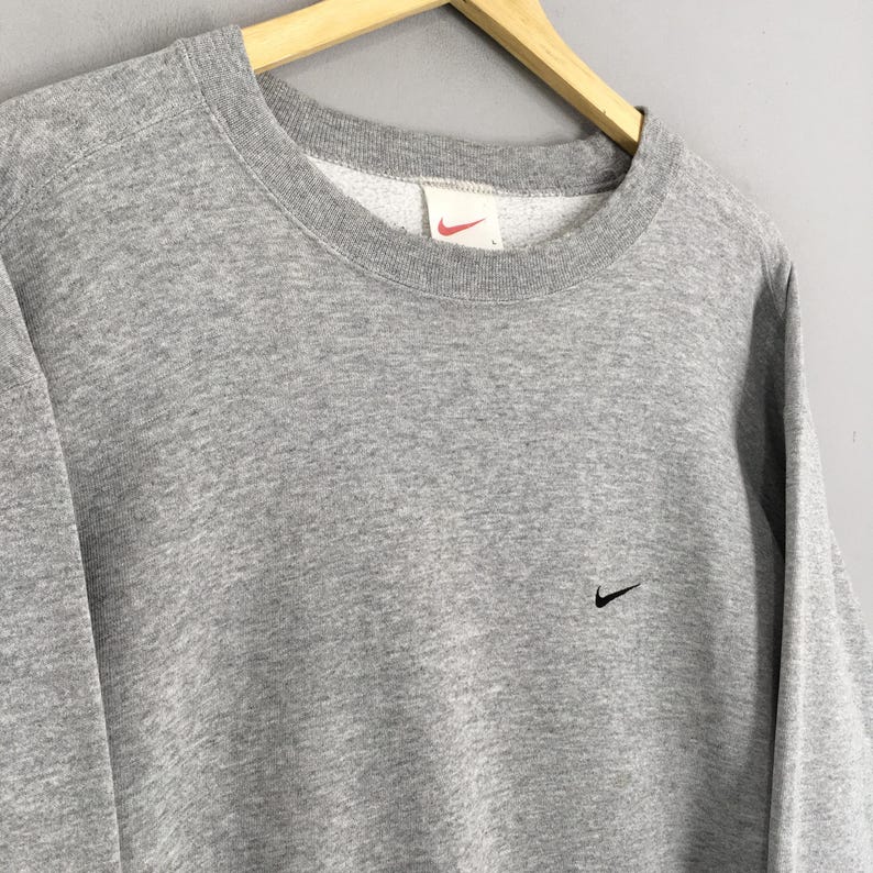 May include: A gray crew neck sweatshirt with a small black Nike swoosh logo on the chest. The sweatshirt has a ribbed neckline and cuffs. The Nike logo is also visible on the tag inside the collar.