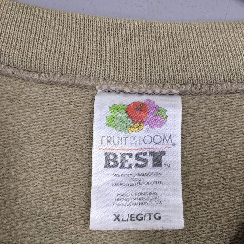 K&ouml;nnte beinhalten: Nahaufnahme eines beigefarbenen Sweatshirts mit einem Fruit of the Loom-Etikett. Das Etikett zeigt das Markenlogo, das Wort "BEST" und Gr&ouml;&szlig;enangaben. Das Sweatshirt besteht aus einer Baumwoll- und Polyestermischung.