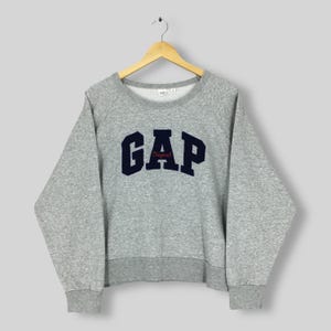 May include: A gray GAP sweatshirt with a crew neck and long sleeves. The word "GAP" is in navy blue block letters across the chest, with "Original" in red script underneath. The sweatshirt is hanging on a wooden hanger.