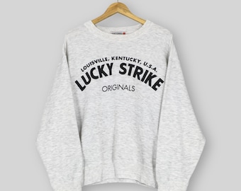 May include: A light grey sweatshirt featuring the black text "LUCKY STRIKE ORIGINALS" arched across the chest. Above this is the text "LOUISVILLE, KENTUCKY, U.S.A.". The sweatshirt is suspended from a wooden hanger.