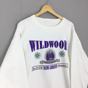 May include: White sweatshirt with the word "WILDWOOD" in purple block letters. Below the word are three circular emblems and the words "WORLD REGATTA" and "NEW JERSEY". The sweatshirt has long sleeves and a crew neck.