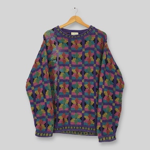 May include: A colourful vintage sweater with a geometric design. The sweater displays a mix of purple, teal, pink, yellow, and black squares. It has a crew neck and long sleeves, hanging on a wooden hanger.