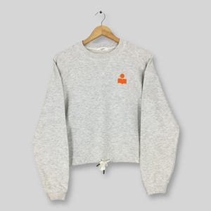 May include: A light gray sweatshirt with long sleeves and a drawstring at the waist. An orange logo is on the upper left chest. The sweatshirt is hanging on a wooden hanger.