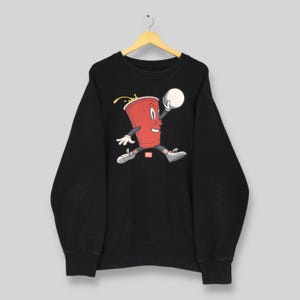 Puede incluir: Sudadera negra con una ilustración de dibujos animados de una taza roja con cara, brazos y piernas, botando una pelota blanca. La taza tiene líquido amarillo que se derrama. La sudadera está colgada de una percha de madera.