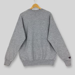May include: A heather gray sweatshirt with long sleeves, a crew neck, and ribbed cuffs and hem. The sweatshirt is hanging on a wooden hanger. A small logo is visible on the left sleeve.