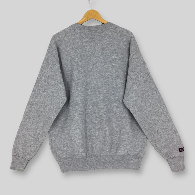 May include: A heather gray sweatshirt with long sleeves, a crew neck, and ribbed cuffs and hem. The sweatshirt is hanging on a wooden hanger. A small logo is visible on the left sleeve.