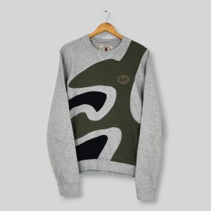 May include: A gray sweatshirt with a unique design. The sweatshirt features a large olive green and black abstract pattern on the front. The sleeves and upper body are gray. A small emblem is on the upper chest.