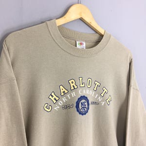 K&ouml;nnte beinhalten: Ein beigefarbenes Sweatshirt mit dem Wort "CHARLOTTE" in gelben Buchstaben, die sich &uuml;ber der Brust w&ouml;lben. Darunter steht der Text "NORTH CAROLINA USA" und ein blaues Emblem. Das Sweatshirt h&auml;ngt an einem Holzb&uuml;gel.