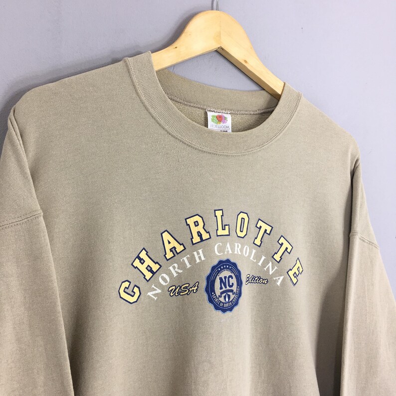 K&ouml;nnte beinhalten: Ein beigefarbenes Sweatshirt mit dem Wort "CHARLOTTE" in gelben Buchstaben, die sich &uuml;ber der Brust w&ouml;lben. Darunter steht der Text "NORTH CAROLINA USA" und ein blaues Emblem. Das Sweatshirt h&auml;ngt an einem Holzb&uuml;gel.