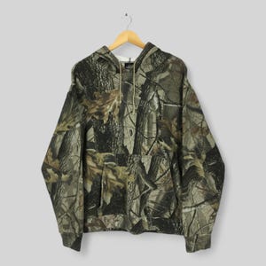Peut inclure: Un sweat &agrave; capuche &agrave; motif camouflage avec une capuche &agrave; cordon et une poche kangourou. Le motif pr&eacute;sente un m&eacute;lange de couleurs marron, vert et beige, ressemblant &agrave; un environnement forestier. Le sweat est suspendu &agrave; un cintre en bois.