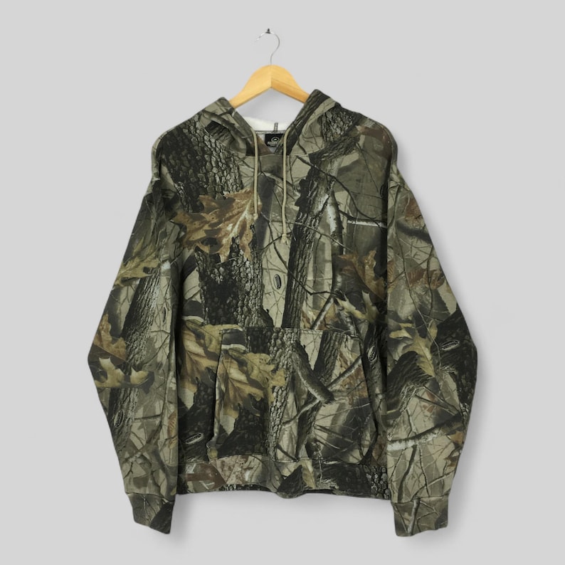 Peut inclure: Un sweat &agrave; capuche &agrave; motif camouflage avec une capuche &agrave; cordon et une poche kangourou. Le motif pr&eacute;sente un m&eacute;lange de couleurs marron, vert et beige, ressemblant &agrave; un environnement forestier. Le sweat est suspendu &agrave; un cintre en bois.
