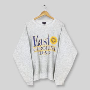 以下が含まれることがあります： 「East Carolina Dad」の文字が青と金色の文字でプリントされた、ライトグレーのスウェットシャツ。テキストの右側には円形のエンブレムがあります。スウェットシャツは木製のハンガーに吊るされています。