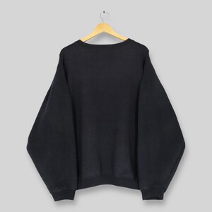 Op de afbeelding: Een zwart sweatshirt met ronde hals hangt aan een houten hanger. De sweatshirt heeft lange mouwen en een relaxte pasvorm. Het kledingstuk is gemaakt van een zachte, donkere stof. De achtergrond is effen lichtgrijs.