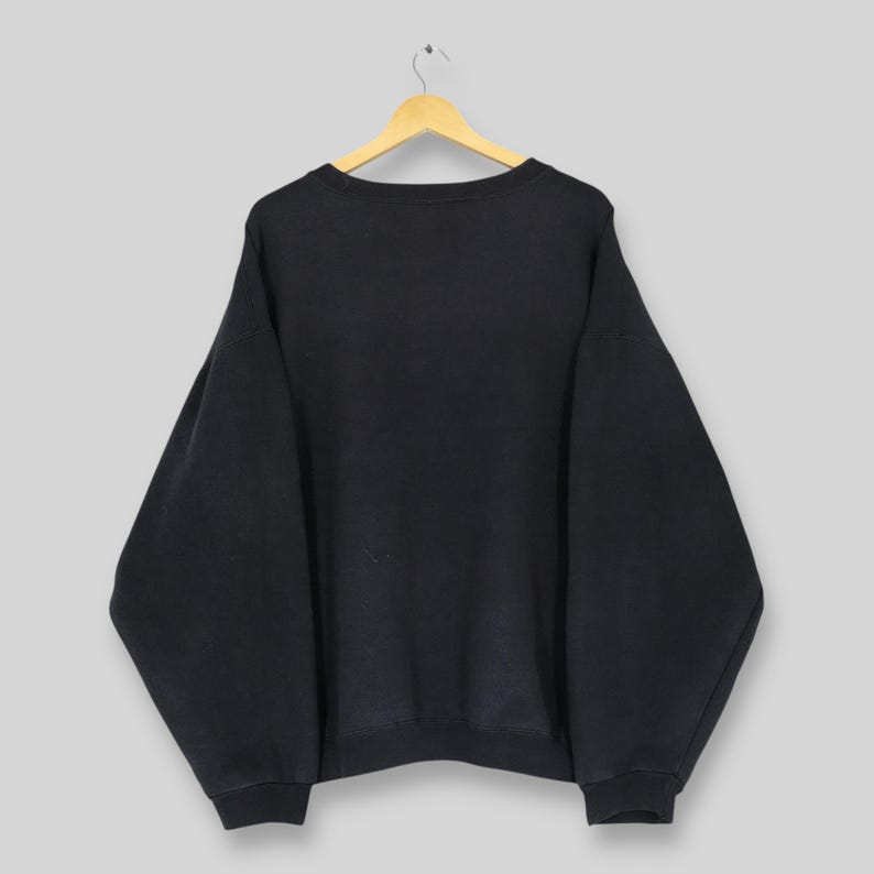 Op de afbeelding: Een zwart sweatshirt met ronde hals hangt aan een houten hanger. De sweatshirt heeft lange mouwen en een relaxte pasvorm. Het kledingstuk is gemaakt van een zachte, donkere stof. De achtergrond is effen lichtgrijs.