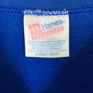 May include: Close-up of a blue garment with a white tag. The tag features the "Hanes ACTIVEWEAR" logo in red and white. Text on the tag indicates the fabric composition and origin. The garment is labeled "X LARGE/ GRAND".