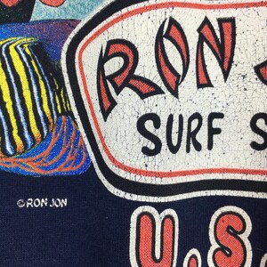 May include: Close-up of a navy blue t-shirt with a distressed graphic. The graphic features the text "RON JON SURF SHOP" in white, red, and black. A colourful fish illustration is also visible.