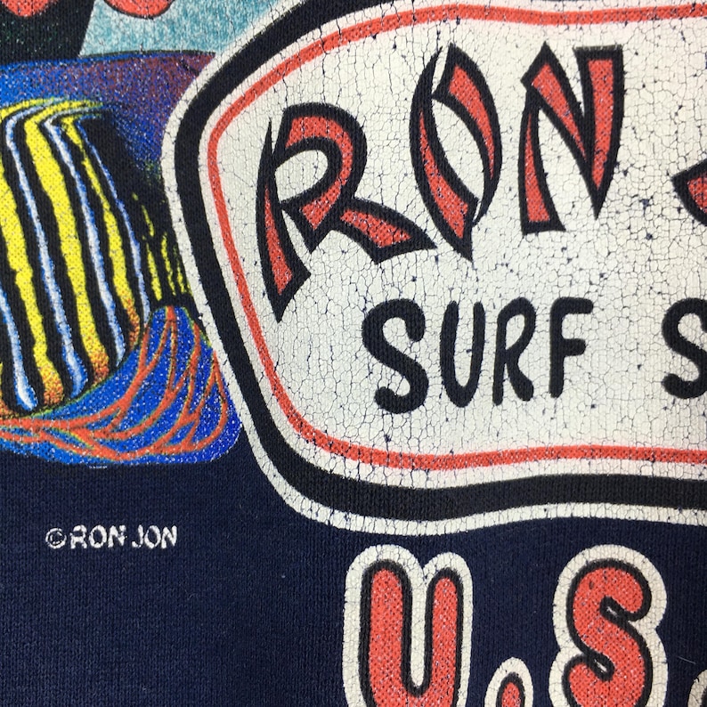 May include: Close-up of a navy blue t-shirt with a distressed graphic. The graphic features the text "RON JON SURF SHOP" in white, red, and black. A colourful fish illustration is also visible.