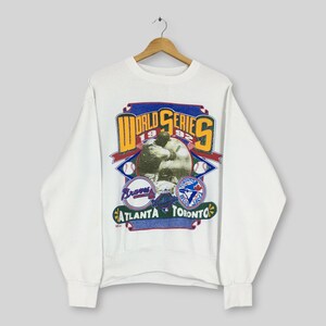 May include: A white long-sleeve sweatshirt featuring a 1992 World Series graphic. The design includes the text World Series, Atlanta, and Toronto, along with a baseball player illustration, two baseballs, and the Atlanta Braves and Toronto Blue Jays logos.