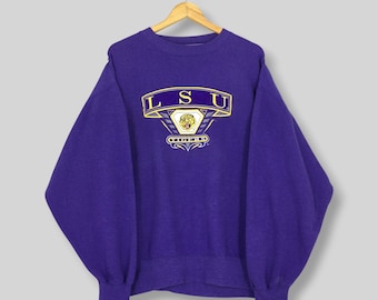 May include: A purple sweatshirt with the letters "LSU" in gold and white, along with a tiger emblem. The sweatshirt is hanging on a wooden hanger. The sleeves are long and the cuffs are ribbed.