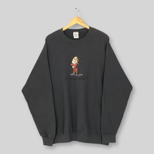 Peut inclure: Sweat-shirt gris foncé avec un dessin animé d'un nain et le texte "Size doesn't matter". Le nain porte une veste rouge et des chaussures marron. Le sweat-shirt est suspendu à un cintre en bois.