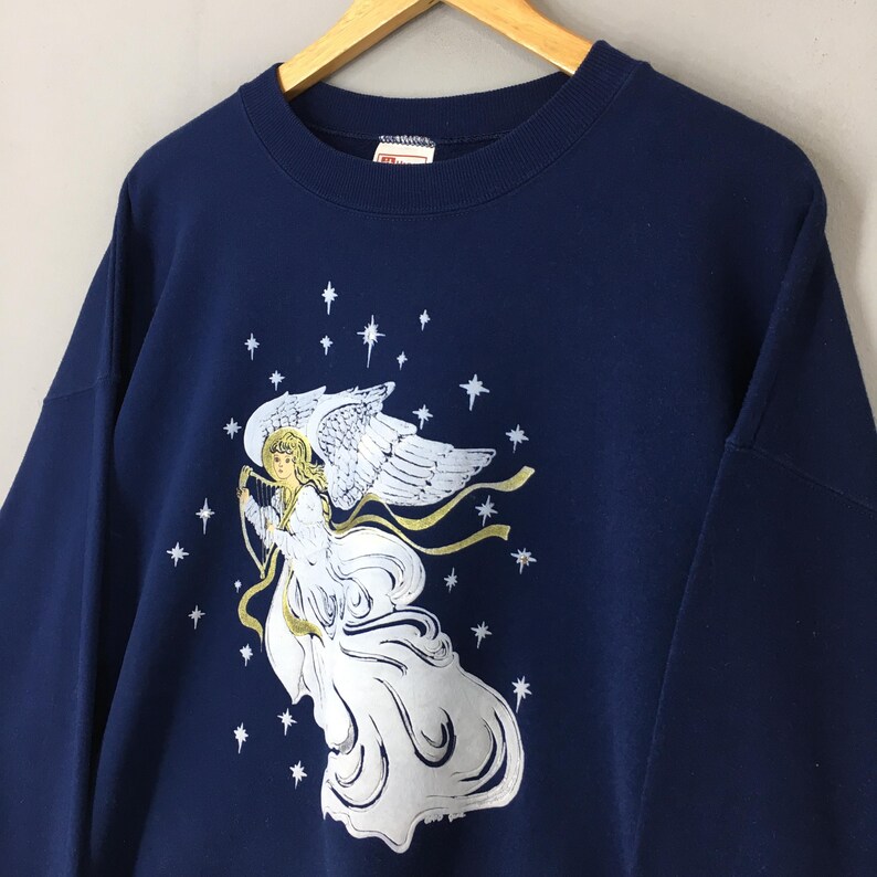 May include: A navy blue long-sleeve sweatshirt featuring a graphic print of an angel with white wings and a flowing white gown, playing a harp. The design is surrounded by white stars and gold ribbons, set against the solid dark blue fabric.
