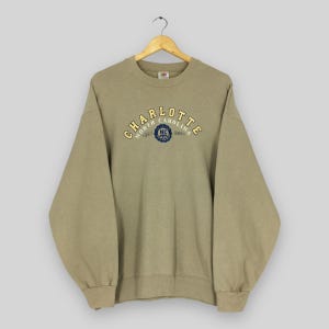 K&ouml;nnte beinhalten: Beige Sweatshirt mit gelbem und blauem Schriftzug "CHARLOTTE NORTH CAROLINA". Das Kleidungsst&uuml;ck h&auml;ngt an einem Holzkleiderb&uuml;gel.